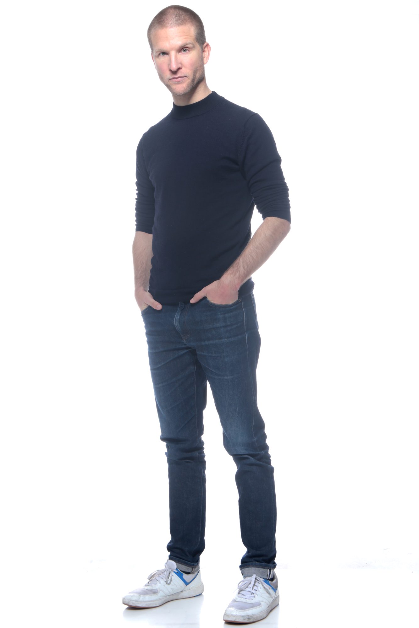 En pied, la main dans la poche, pull noir, jean et baskets