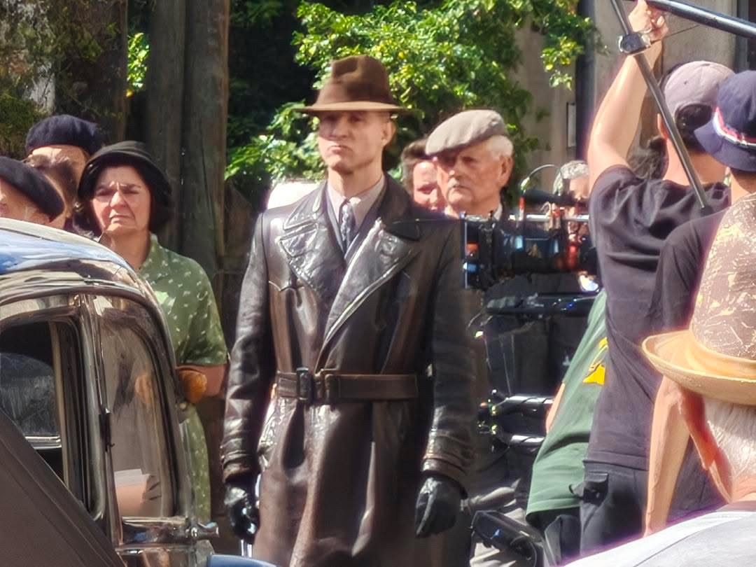 Sur un tournage, en manteau de cuir et chapeau, devant une voiture d'époque