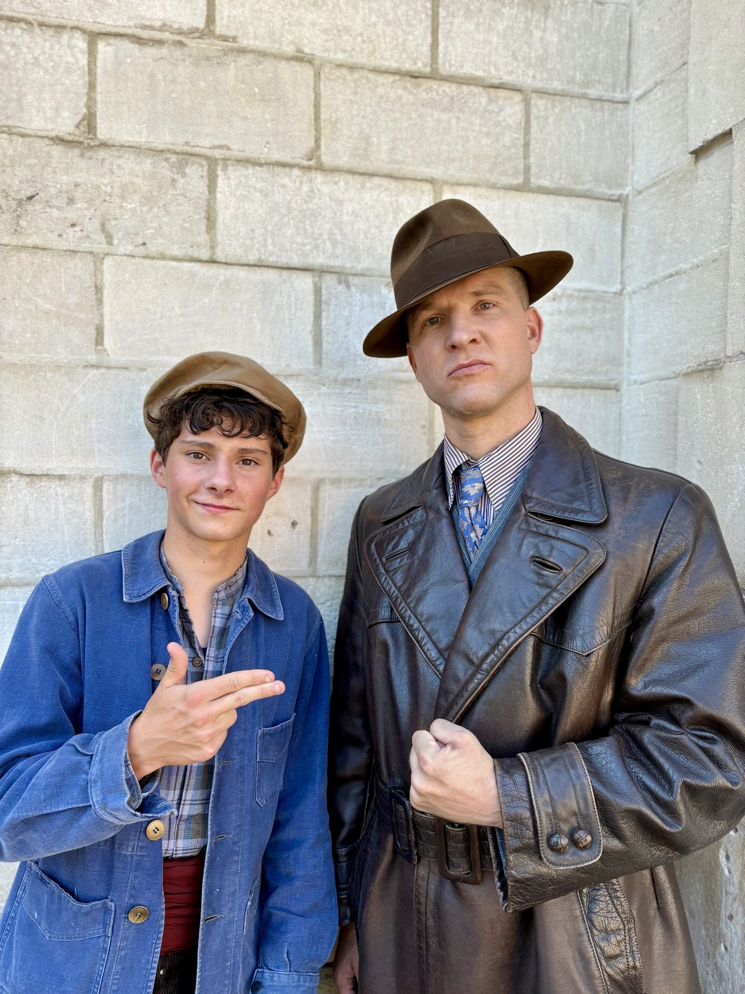 En manteau de cuir et chapeau, avec un jeune comédien, devant un mur de pierre