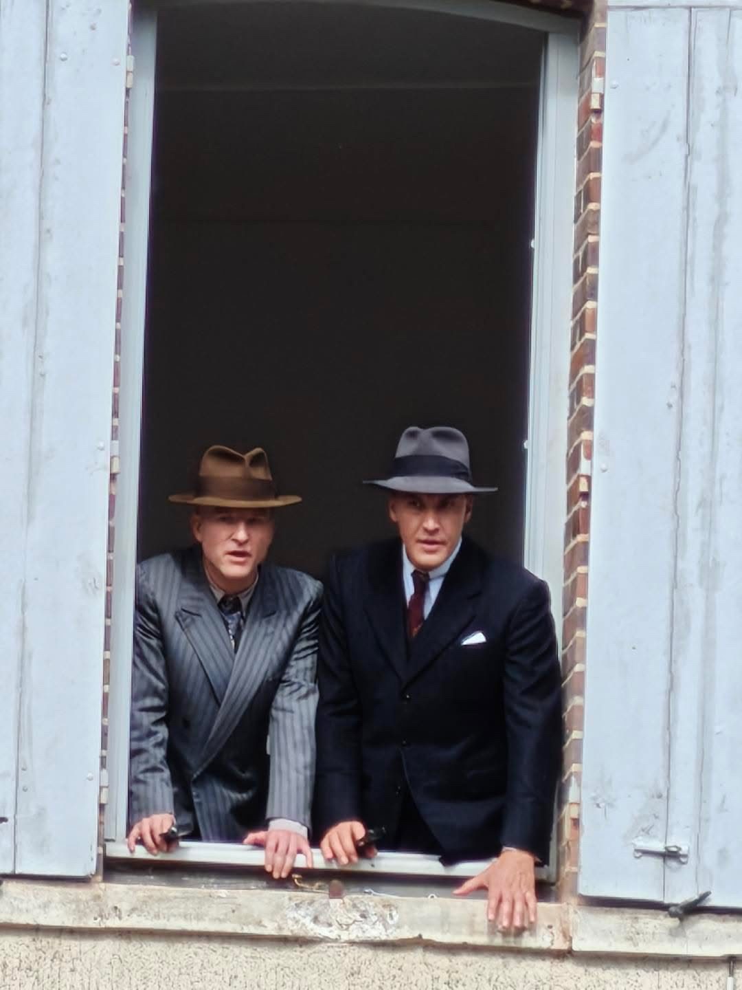 Deux hommes en costume et chapeau penchés à une fenêtre aux volets bleus